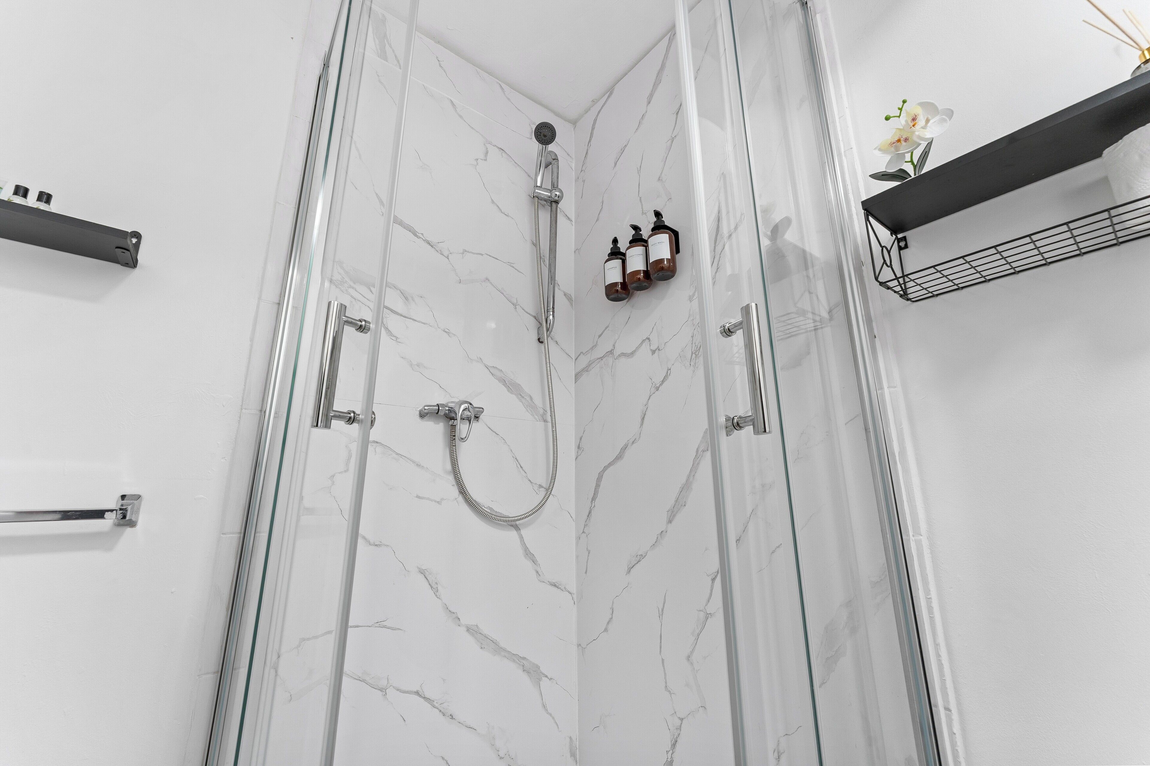 A modern bathroom with a glass shower enclosure, marble walls, and a black shelf with toiletries.