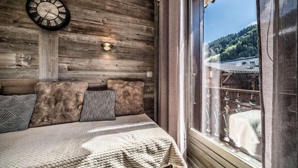 A cozy room with a wooden wall, a bed with a knitted blanket, and a view of a forested area through a window.