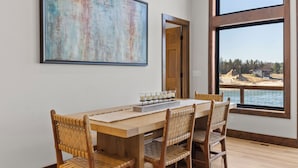 A dining area with a wooden table and chairs, a large abstract painting, and a view of a beach and forest through large windows. - Lake House for 14! Fire Pit + Game Room (New Lisbon)