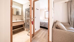 A modern room with a wooden floor, a beige sofa, a wooden-framed door, a bathroom with a sink and towel rack, and a bed with a gray blanket. - 140m from ski lifts, 1st floor, balcony, tv, 33m², Morzine (Morzine)