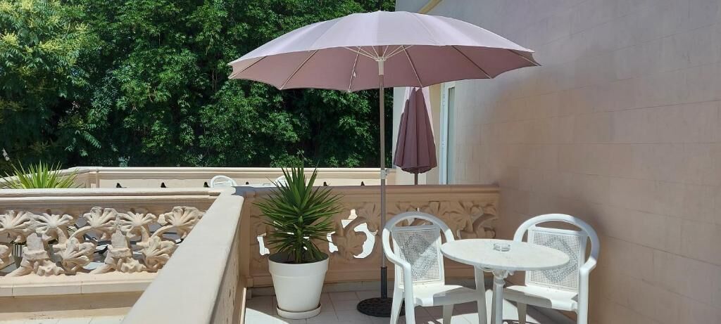 Un balcón con una mesa y sillas blancas, una planta en maceta y un paraguas púrpura.