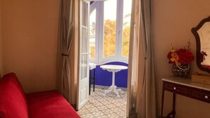 A room with a red sofa, a small white table, a mirror, and a view of the outside through a window with curtains. - Hotel Noucentista (Sitges)
