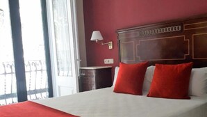 A hotel room with a large bed, red pillows, a wooden headboard, a nightstand, and a window with a view. - Hotel Noucentista (Sitges)