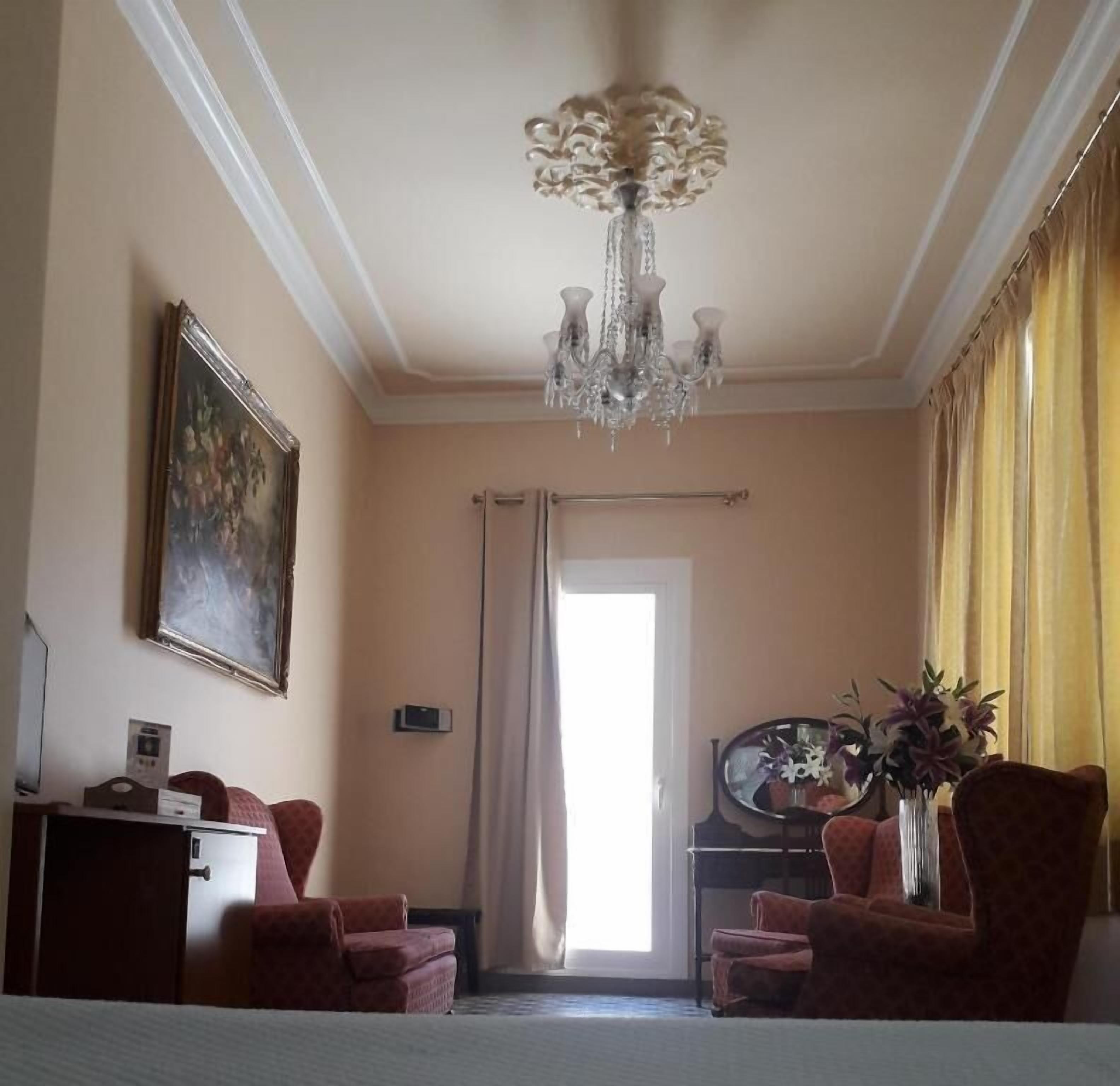 A hotel room with a chandelier, a painting, two armchairs, a desk, a mirror, and a window with curtains.