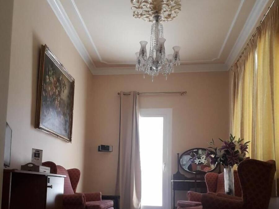 Habitación de hotel con candelabro, un cuadro, dos sillones, un escritorio, un espejo y una ventana con cortinas.