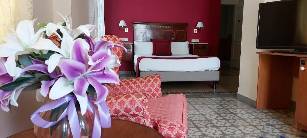 A hotel room with a bed, a TV, a vase with flowers, and a red wall.