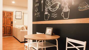 Un rincón comedor acogedor con una pared de pizarra decorada con dibujos hechos a tiza de comida y utensilios, una mesa redonda de madera y sillas blancas.