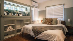 Un dormitorio con una cama, un tocador con espejo, repisas con toallas y una ventana con persianas.