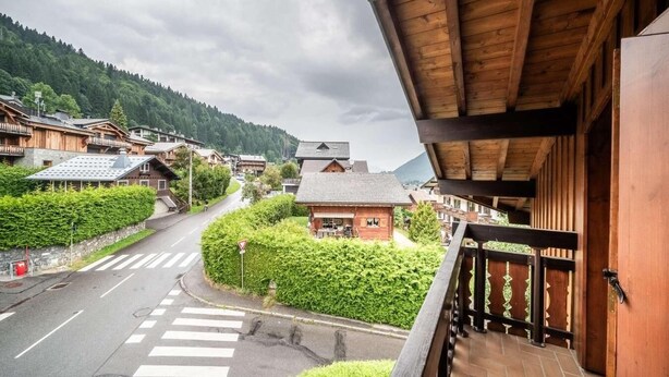 Un bâtiment en bois doté d’un balcon donnant sur une route, ainsi qu’une petite maison entourée de verdure.