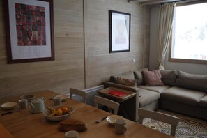 A cozy living room with a wooden wall, a sofa, a dining table with a bowl of fruit, and framed pictures.