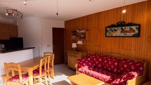 A cozy living room with a wooden sofa, matching chairs, a wooden coffee table, and a kitchen area with wooden cabinets and a hanging light fixture.