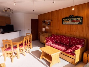 Uma sala de estar acolhedora com um sofá de madeira, poltronas combinando, uma mesa de centro de madeira e um espaço de cozinha com armários de madeira e um lustre.