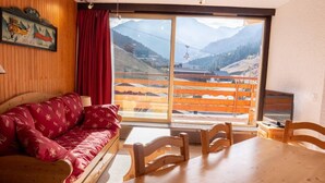 A room with a wooden sofa, a dining table, and chairs. A large window offers a view of mountains and a ski lift.