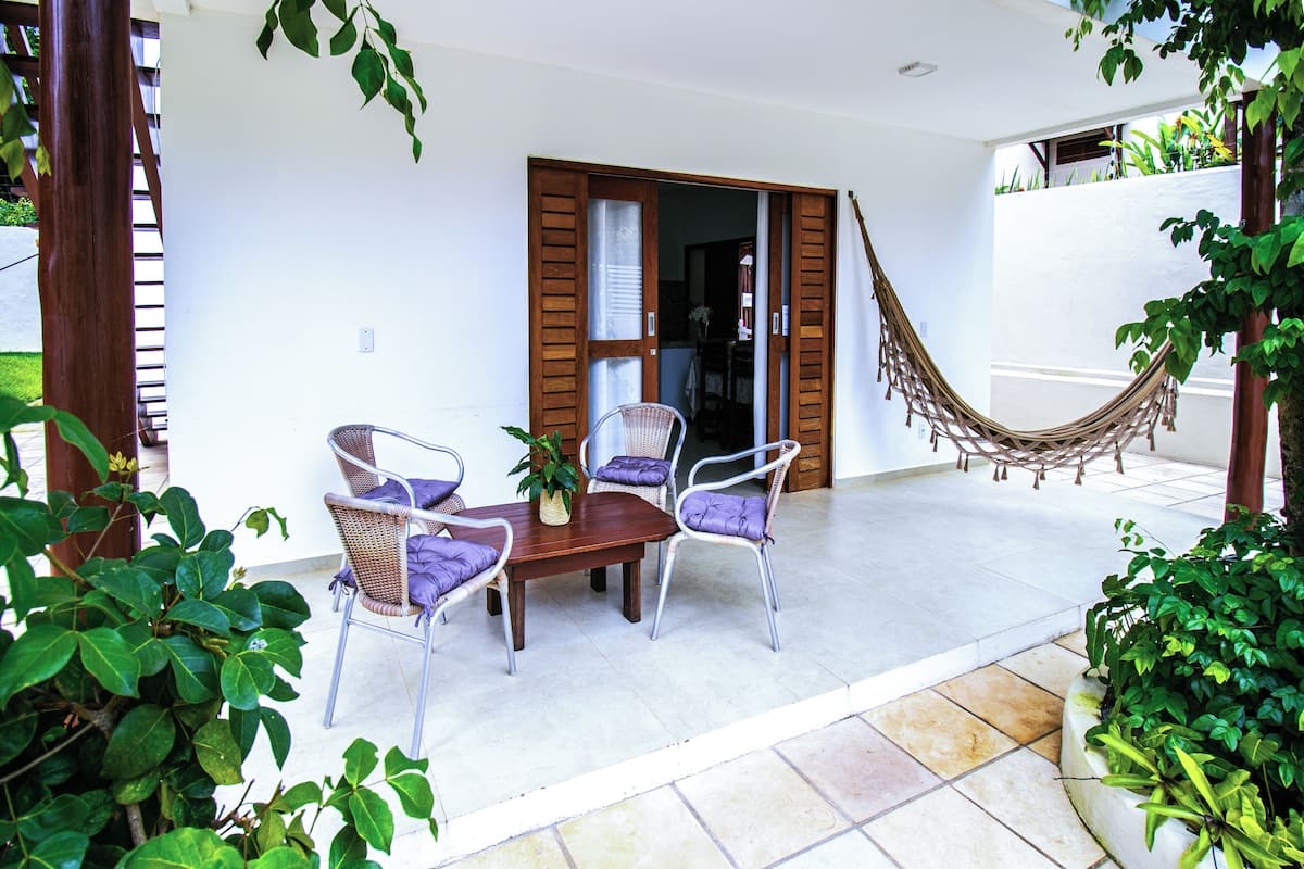 An outdoor patio with a wooden table and wicker chairs, a hammock, and potted plants.