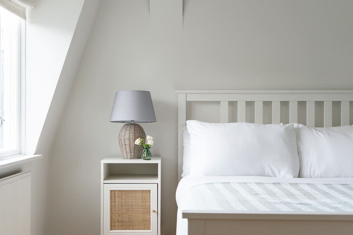 Une chambre à coucher avec un lit blanc, une table de chevet avec une lampe et un petit vase avec des fleurs.