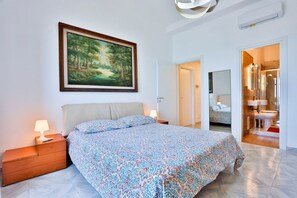 Une chambre à coucher avec un lit, des tables de chevet, un tableau, une salle de bain visible à travers une porte ouverte et un sol carrelé.