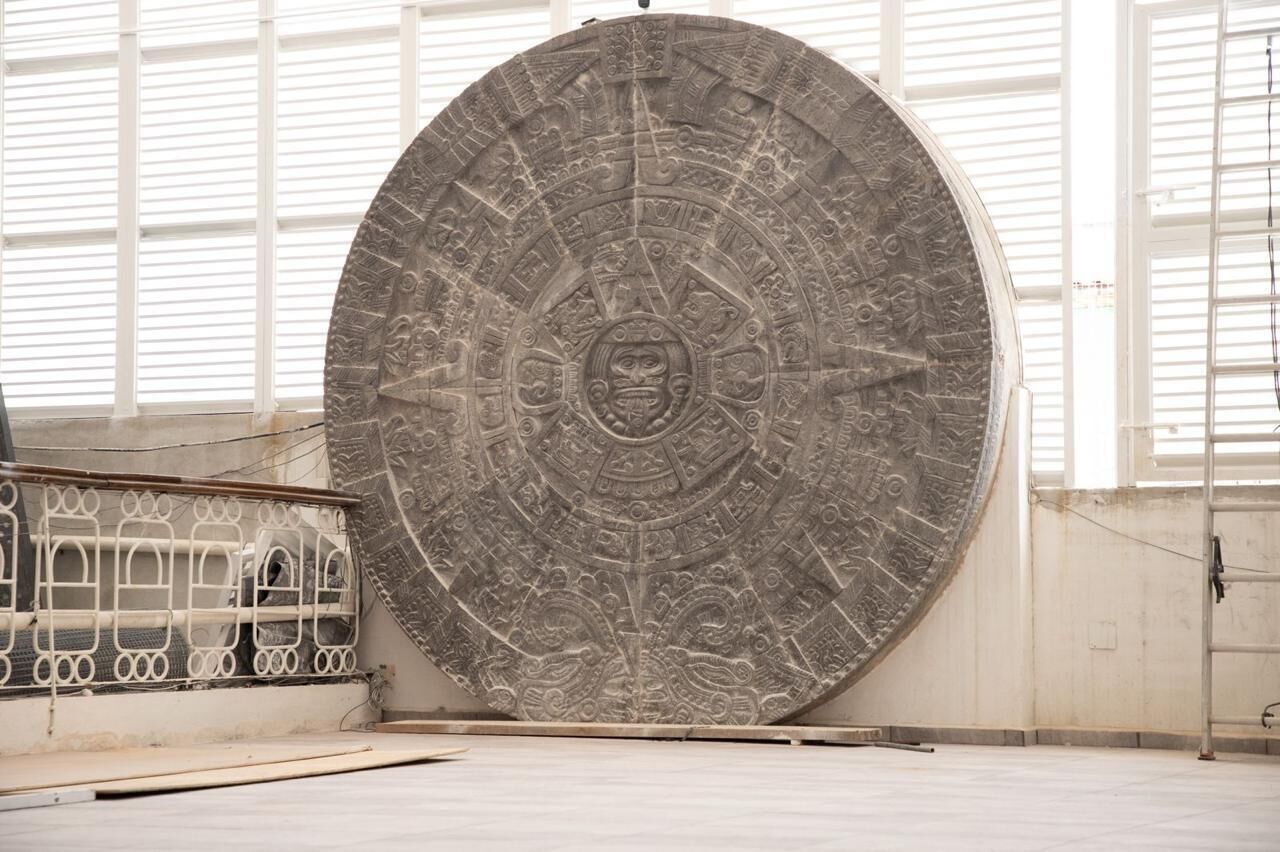 Un grand artefact circulaire en pierre, orné de sculptures complexes, est exposé à l’intérieur, avec une rambarde en métal et de grandes fenêtres en arrière-plan.