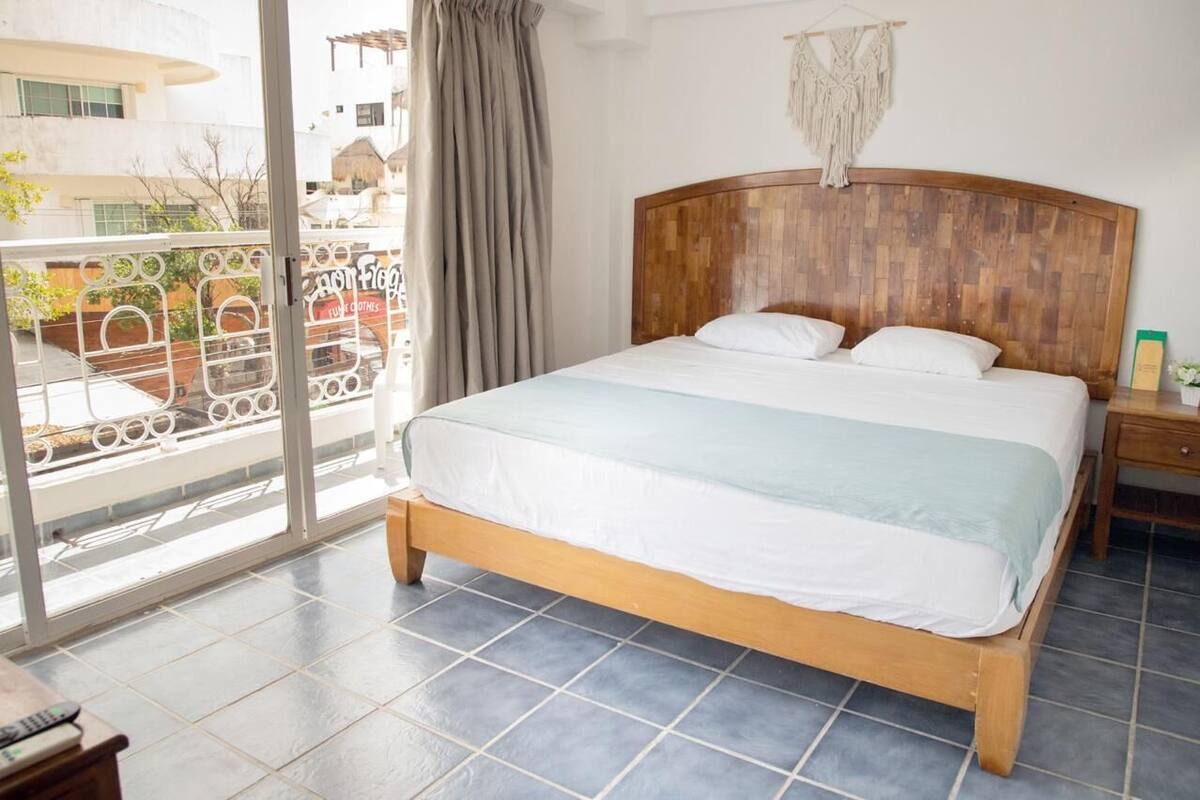Une chambre à coucher avec un lit en bois, une table de chevet, une fenêtre donnant sur des immeubles et un rideau.