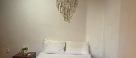 A neatly made bed with white linens and a green bedspread, a bedside table with a plant, and a macrame wall hanging.