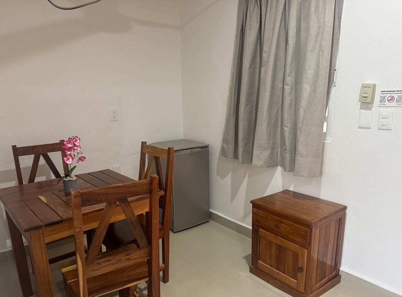 A small room with a wooden table, chairs, a small cabinet, a refrigerator, and a window with curtains.