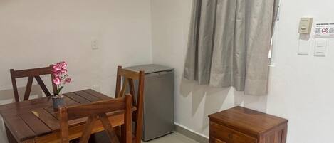 A small room with a wooden table, chairs, a small cabinet, a refrigerator, and a window with curtains.
