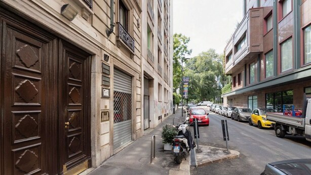 Une rue étroite en milieu urbain, bordée de voitures garées, avec une moto et un bâtiment doté de grandes portes en bois et d’un portail en métal.
