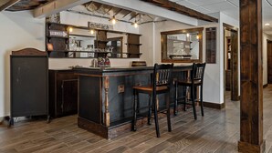 Un espace bar avec un comptoir en bois foncé, trois tabourets de bar et des étagères remplies de verres et de bouteilles.