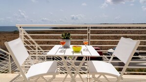 Una mesa metálica blanca con dos sillas, una planta en maceta, un cuenco y dos vasos de líquido anaranjado, con el mar de fondo.