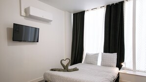 A neatly made bed with a geometric patterned bedspread, a heart-shaped towel arrangement, a flat-screen TV mounted on the wall, and a window with black curtains. - Le Pied-à-Terre - Wifi - Centre-ville (Laval)