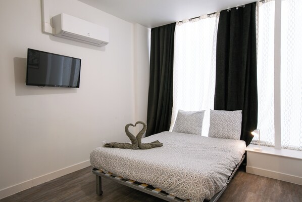 A neatly made bed with a geometric patterned bedspread, a heart-shaped towel arrangement, a flat-screen TV mounted on the wall, and a window with black curtains.