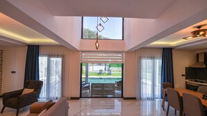 A modern living room with a sofa, armchair, dining table, and chairs. There is a large window with a view of a pool and outdoor area.