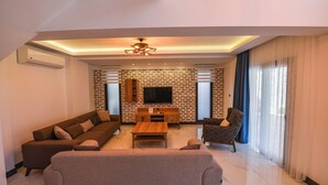 A modern living room with a sofa, armchair, and coffee table. There is a TV mounted on a brick wall and a ceiling light fixture.