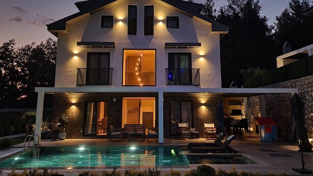 A two-story house with a swimming pool, outdoor seating, and a well-lit entrance.