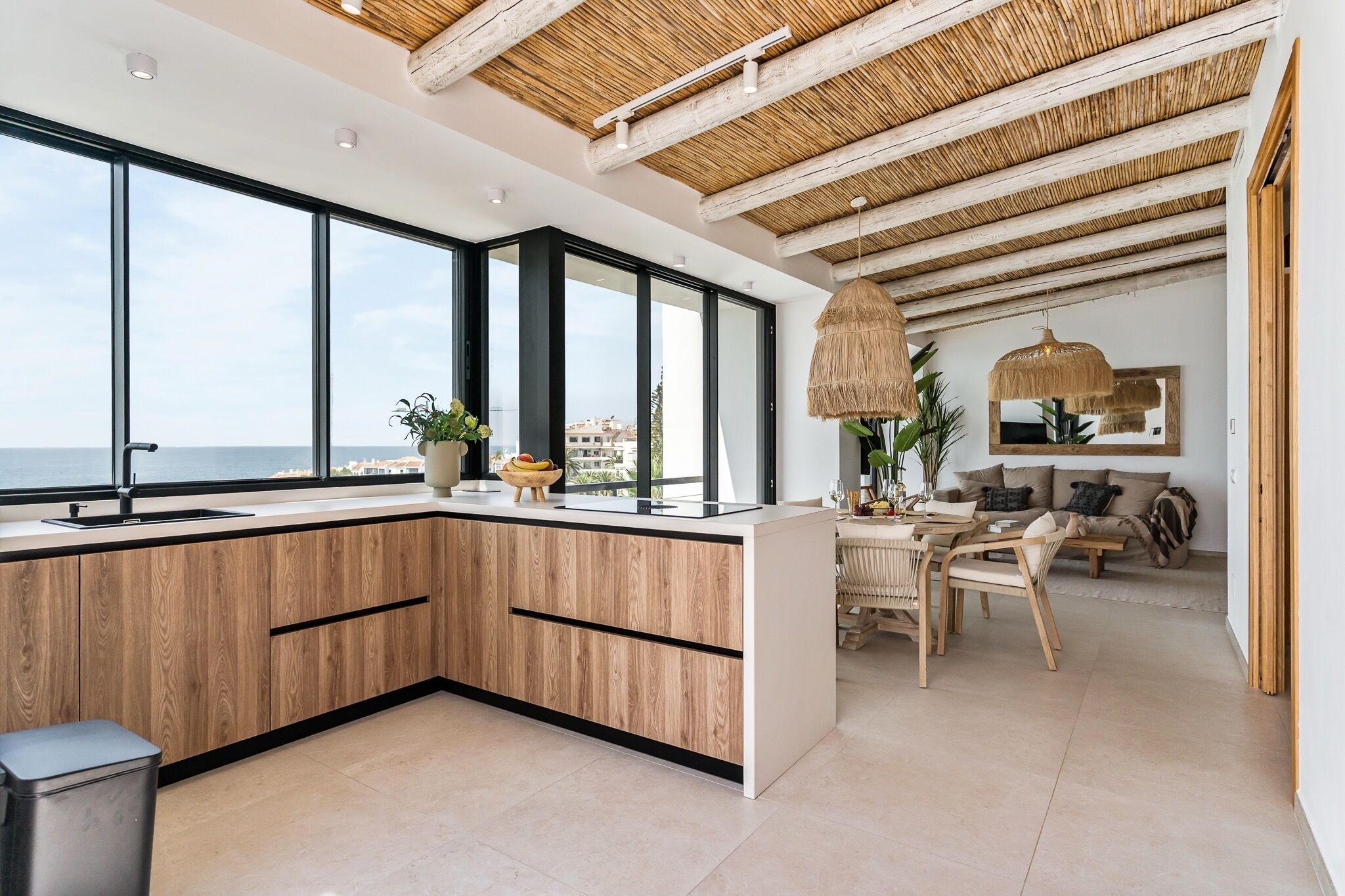 A modern kitchen with large windows, a dining area with a table and chairs, and a view of the sea.