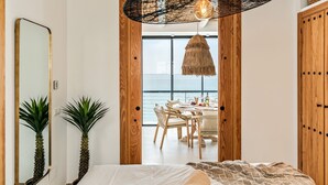 Ein Schlafzimmer mit einem großen Bett, einem Spiegel, einer Topfpflanze und Blick auf einen Essbereich mit Tisch und Stühlen.