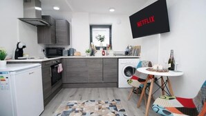 Uma cozinha compacta com micro-ondas, torradeira e pia. Há uma máquina de lavar roupa e uma área de refeições com mesa e cadeiras. Uma TV na parede exibe o logotipo da Netflix.