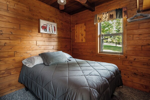A wooden cabin room with a bed, a window with curtains, and a poster on the wall.