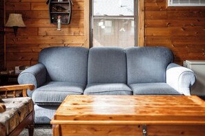 A wooden interior with a grey sofa, a wooden coffee table, a lamp, and a shelf with decorative items.