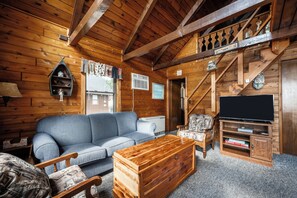 Interior acogedor de una cabaña con techo de madera, un sofá, una mesa de centro, un mueble para televisión y una escalera.