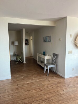 Amplia sala de estar con piso de madera, una mesa consola blanca, una silla gris y una lámpara.