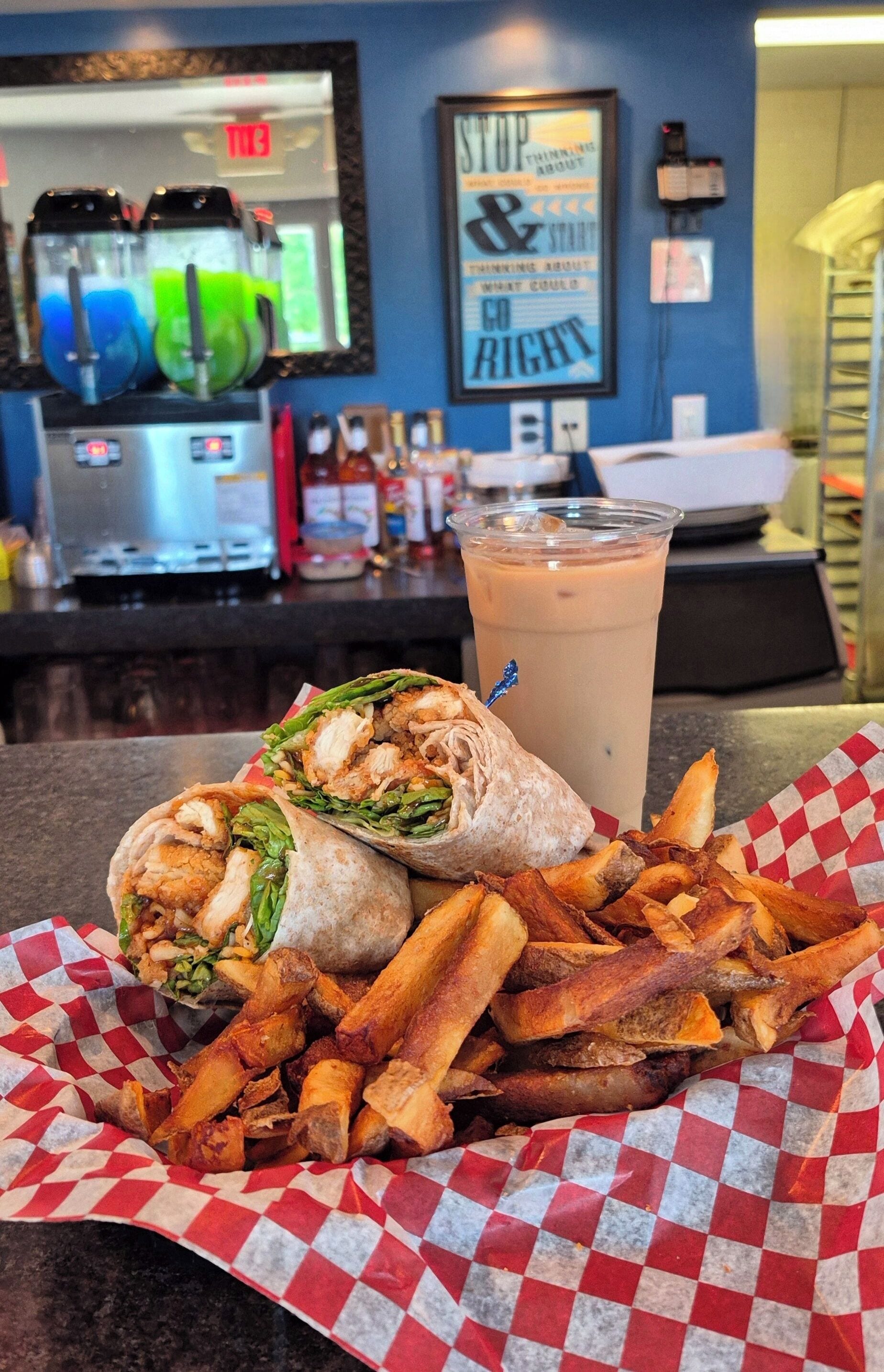 Un plato con un wrap de pollo, papas fritas y una bebida, con un mostrador y equipo de cocina al fondo.