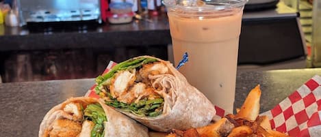 Un repas composé d’un sandwich au poulet, de frites et d’une boisson, avec un comptoir et du matériel de cuisine en arrière-plan.