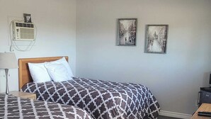 A hotel room with a bed, a bedside lamp, two framed pictures on the wall, and a small table with a remote control. - LaniKai Hotel & Restaurant (Batchewana Bay)