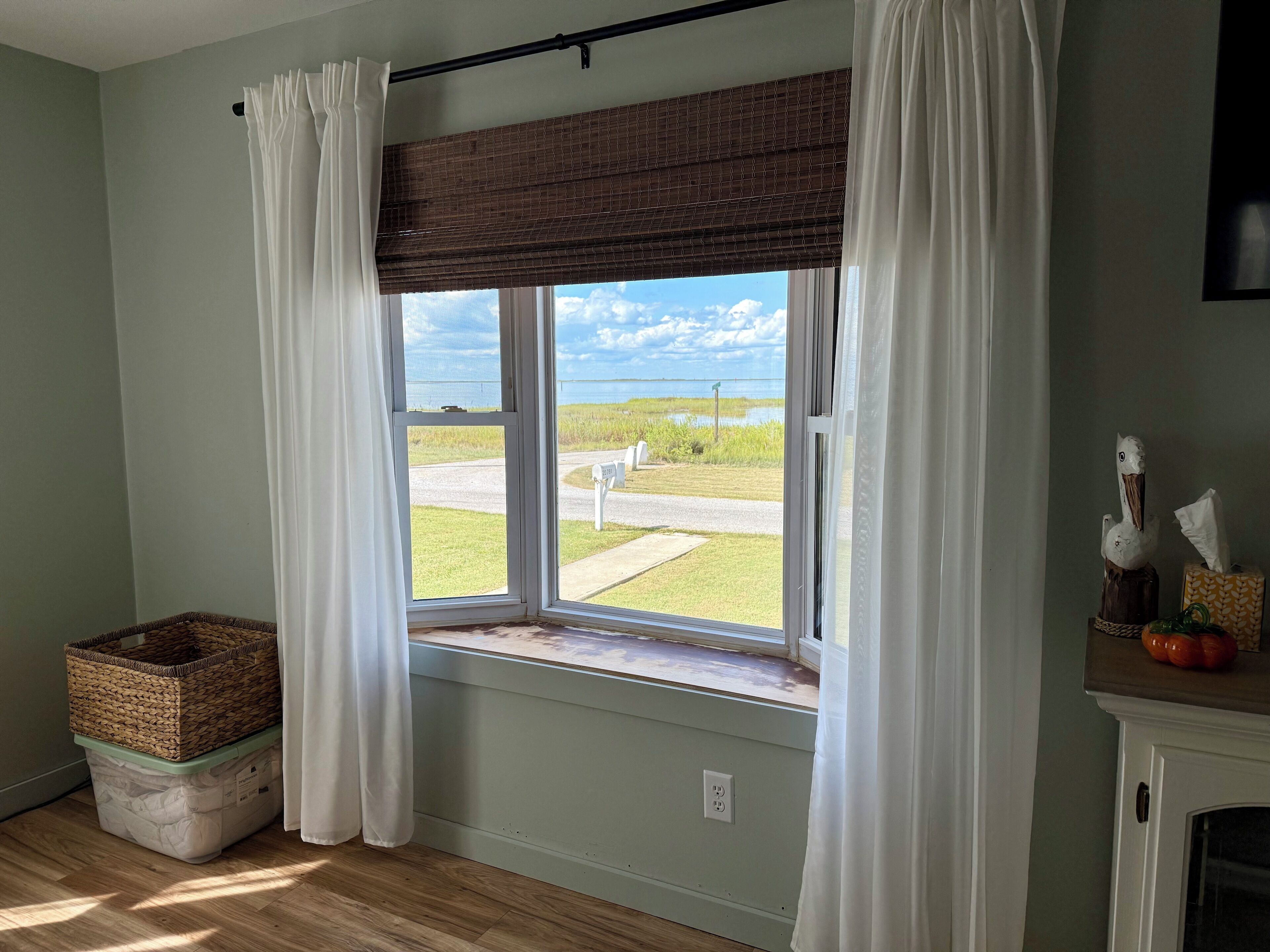 A room with a large window, a wicker basket, a white trash bin, and a small table with a tissue box and decorative items.