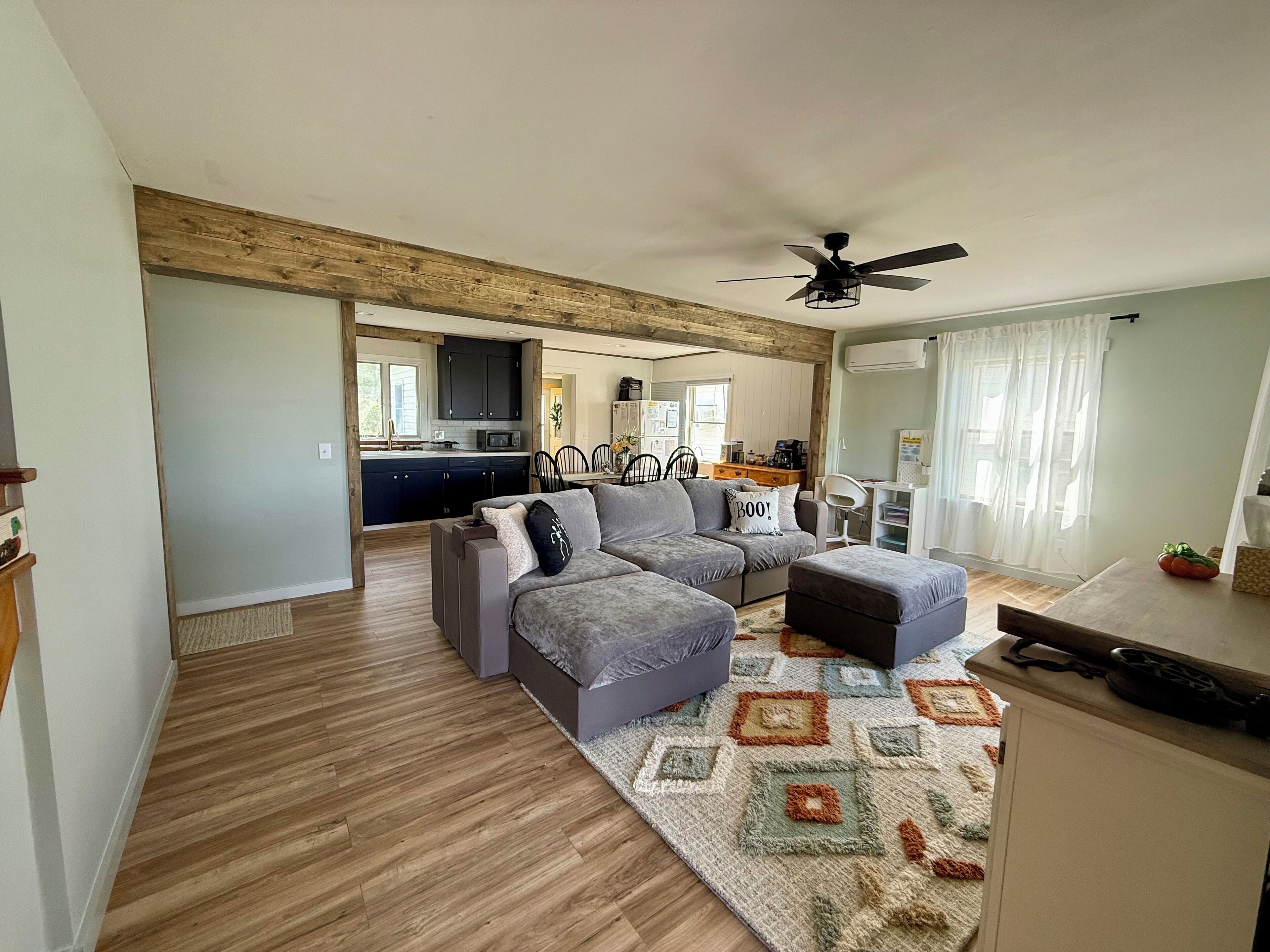 A modern living room with a grey sectional sofa, a small ottoman, and a patterned rug. A ceiling fan is visible, and the room has wooden flooring.
