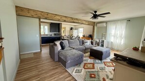 A modern living room with a grey sectional sofa, a small ottoman, and a patterned rug. A ceiling fan is visible, and the room has wooden flooring.