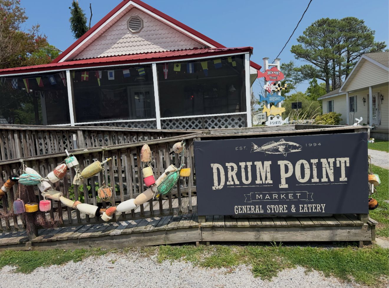 A sign for Drum Point Market with a general store and eatery, a wooden fence, and a red-roofed building in the background.