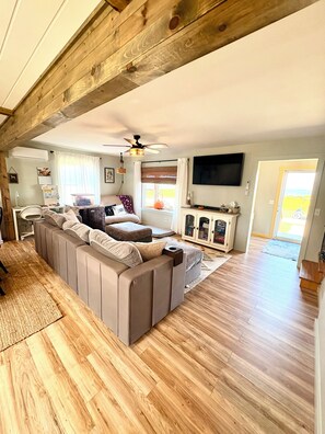 A spacious living room with a large sectional sofa, a ceiling fan, a flat-screen TV, and a wooden floor. - Charming 2 Bedroom house in beautiful Smith Island Pet-Friendly (Smith Island)