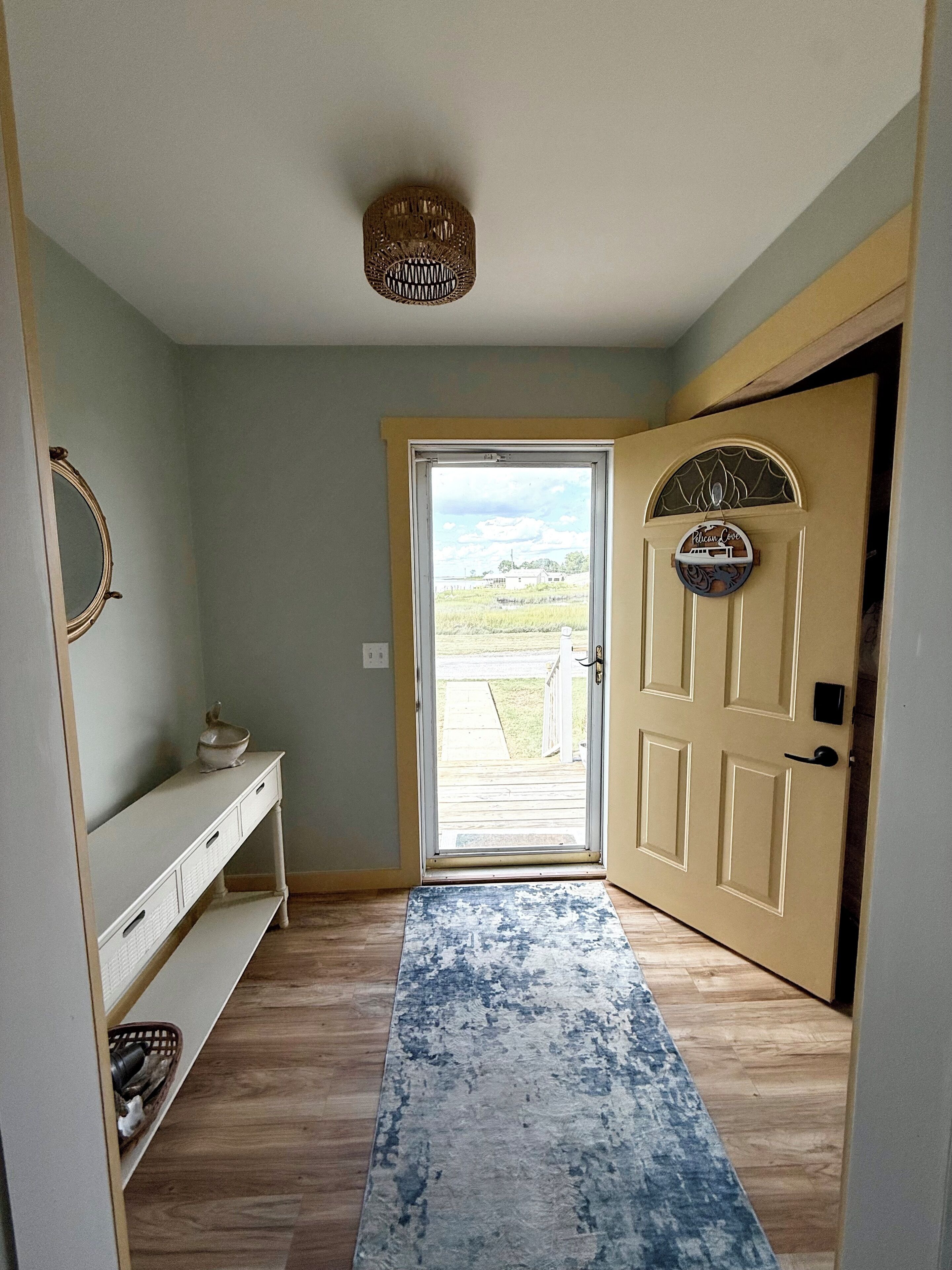 A hallway with a wooden floor, a white bench, a door with a decorative plate, and a view of the outdoors.