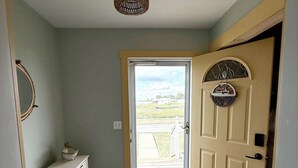 A hallway with a wooden floor, a white bench, a door with a decorative plate, and a view of the outdoors.